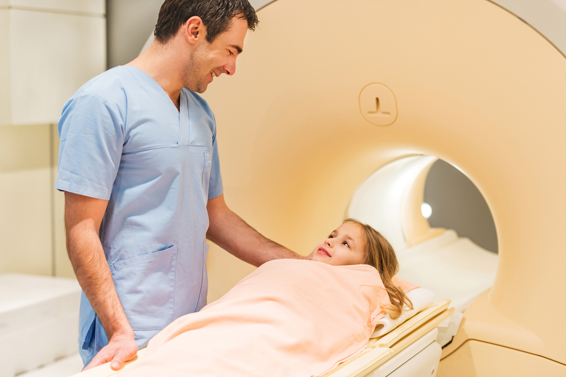 Mid adult radiologist standing next to a little girl who is about to receive an MRI Scan and communicating with her.